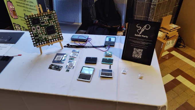 A table with the 10x version of the RP2350 Stamp on it, under it there's the real RP2350 Stamp, Stamp XL, and the Stamp Carrier XL. There is also a photo frame with some information about the company and a QR code to the linktr.ee. There are also multiple devices with a handheld qwerty keyboard. One with trackpad, one without. One with screen and antenna. One with just a large screen. And in the back there seems to be a premium device with a large screen, keyboard, and a case. Connected to a power bank. 