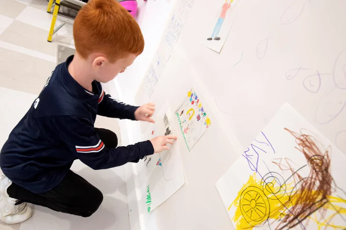 A child wearing a black top and trousers kneeling on the floor adding to a large collage.