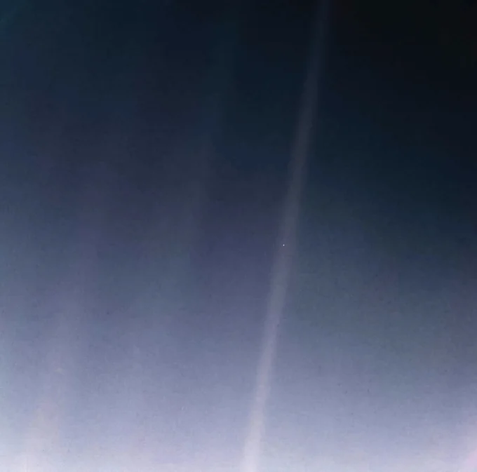 A dark, nearly black background with faint streaks of sunlight forming diagonal beams. In one of these beams, near the right side of the image, a minuscule, barely visible pale blue dot—Earth—stands out, appearing fragile and isolated against the vast emptiness of space. Taken on Feb. 14, 1990, by NASA's Voyager 1 spacecraft from 3.7 billion miles (6 billion kilometers) away, this iconic photograph underscores the profound vastness of the cosmos and Earth's tiny presence within it