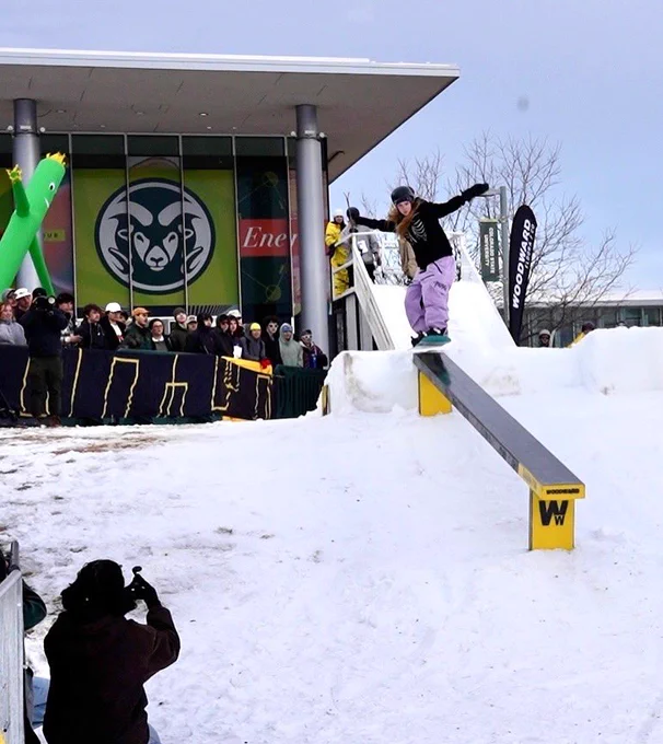 A snowboarder is in midair about to land on a ski rail. A group of students is visible in the image, watching the event
