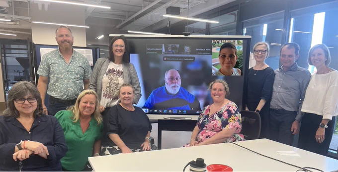 An image of 11 people smiling at the camera. Two of the people are on a zoom screen, whilst the others are seated or standing in a University meeting room.