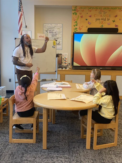 Teacher stands at rhe front of the room while one student raises her hand.