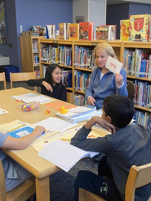 Students sit at a table with a teacher who is holding up a card that says are.
