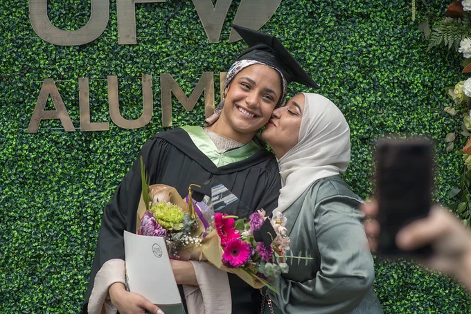A graduate wearing a cap and gown holds flowers while receiving a kiss on the cheek from another woman.