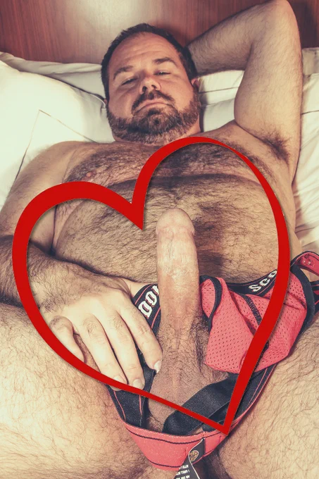 Corbin Sautereau MOCubHot4U lying nude on a bed with an erection. Corbin's penis and face are visible. Corbin's left arm is cocked behind his head, exposing his hairy armpit. Corbin's right hand is gently gripping the base of his penis, and there is a red Double Scorpio jockstrap draped around it as well. There is a red heart outline edited into the photo to surround his erection.