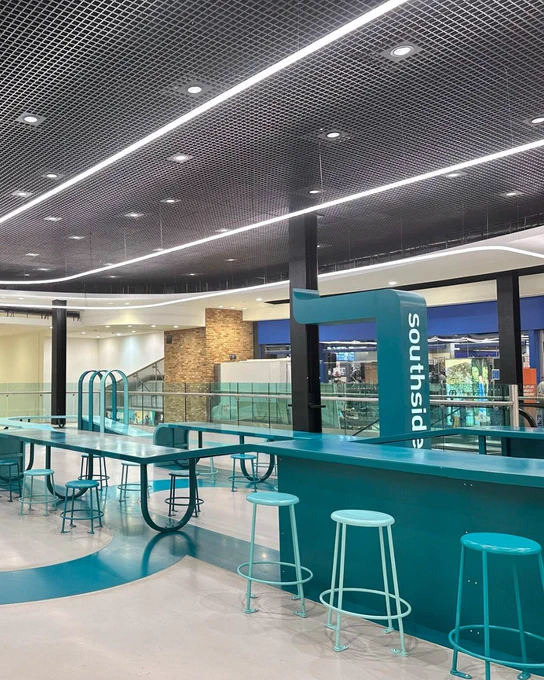 Interior of a seating area with high stools and a bar counter, featuring a prominent Southside logo. The café is under a geometric ceiling light design, and overlooks an indoor area through large windows.