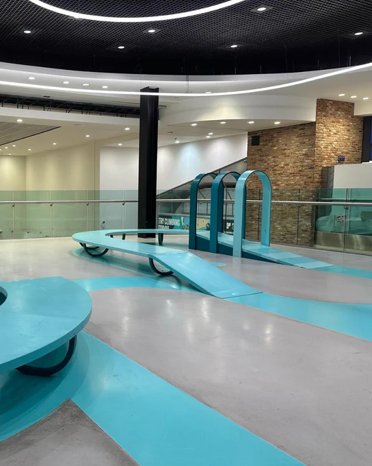 Interior of Southside Wansworth featuring sleek turquoise pathways and modern architectural elements, leading to a spacious, brightly-lit area with a glass barrier and a visible historic brick structure in the background.