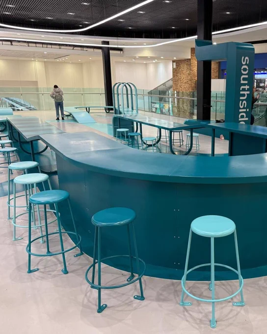 Interior of a modern public space with aqua-colored high stools lined up along a curved counter. In the background, a person stands near the counter opposite the signage "southside".