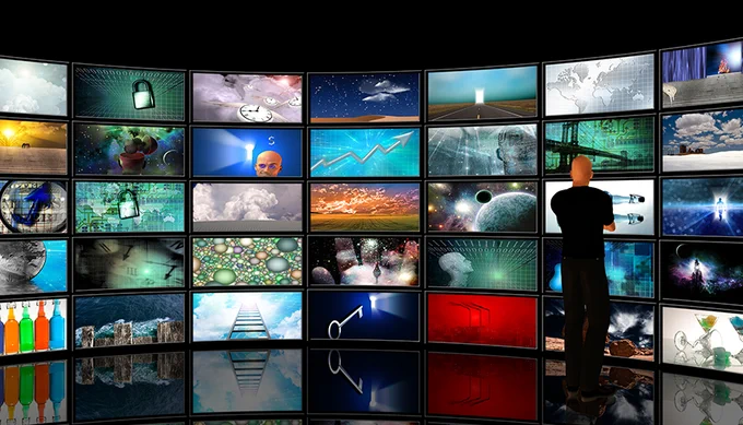 Hombre de espaldas mirando un muro de monitores de televisión, todos ellos con imágenes distintas.