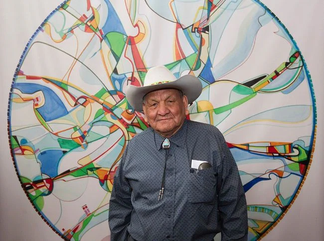 The photo shows an elderly man wearing a white cowboy hat, a blue patterned shirt, and a bolo tie with a turquoise slide. He stands in front of a vibrant, abstract circular painting featuring colorful lines and shapes in blue, green, red, and yellow against a white background. The man has a name tag on his shirt and appears to be at an event or exhibition.