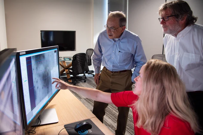 Two men stand behind a woman seated at a computer. She is pointing at a microscopic image on the screen as they look on.