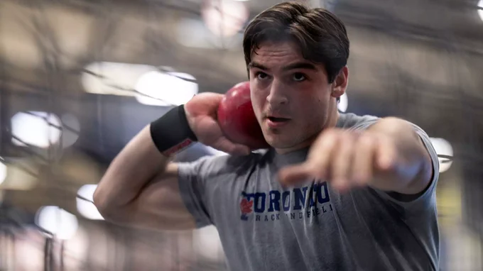 Luka Stoikos holds a shotput ball to his neck while holding his left arm out, preparing to throw it during a University of Toronto Varsity Blues track and field event