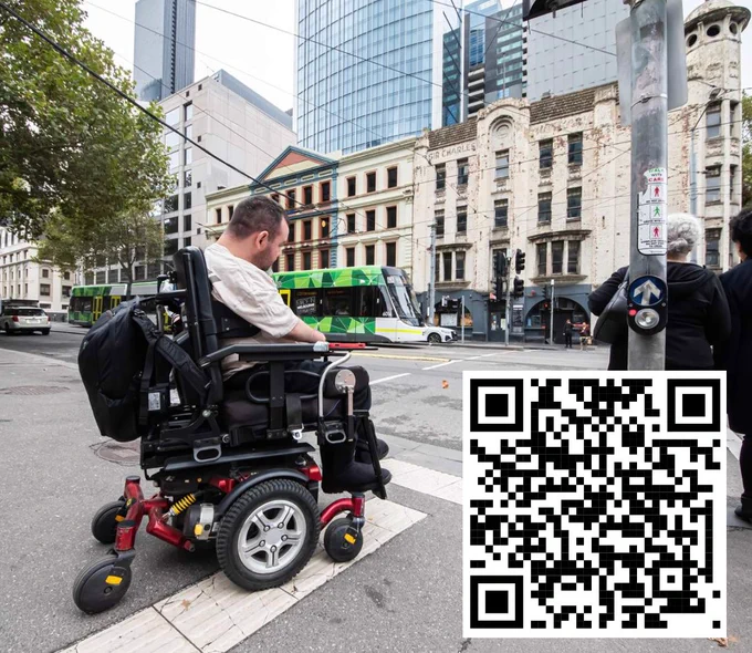 A person with short dark hair seated in a power wheelchair at a pedestrian crossing in the Melbourne CBD with a QR code linking to the ARATA online shop, the URL for which is https://www.arata.org.au/education-resources/online-shop/