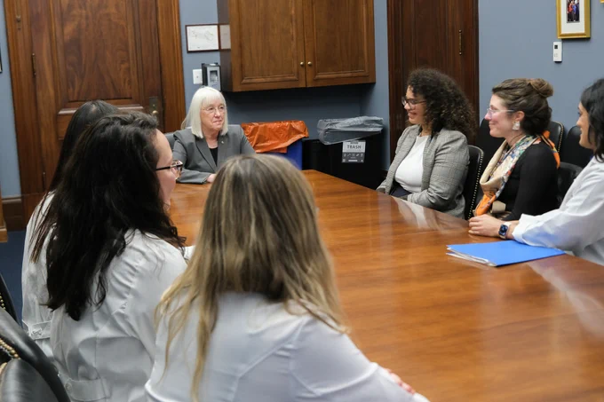 Senator Murray is sitting at the head of a large brown table. Sitting at the table with her are three women in white lab coats on the left and three women, two in business clothes and one in a white lab coat, on the right. The woman on the right in the lab coat is speaking with arm on a blue folder, and the senator is looking at her and smiling.