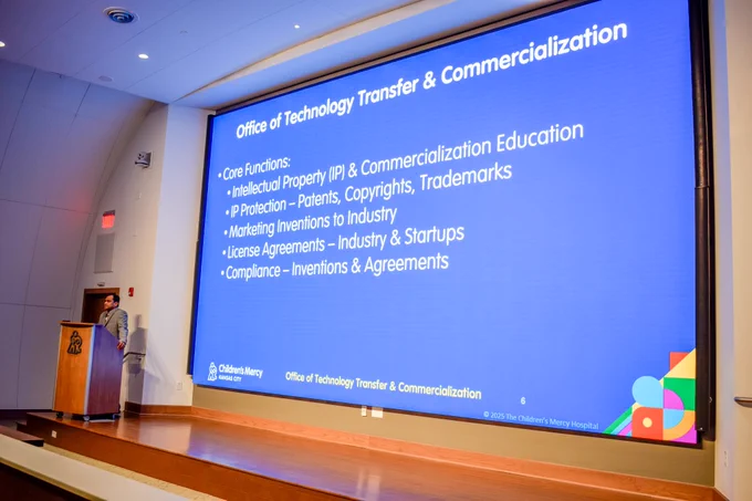 Dr. Aswini Betha stands on a stage at a podium. On the large screen behind him is a slide with a blue background that says "Office of Technology Transfer & Commercialization" and details their core functions. 