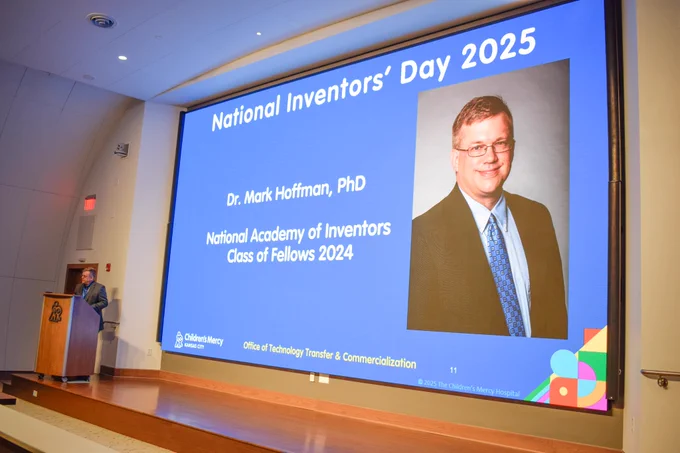 Dr. Mark Hoffman stands on a stage at a podium. On the large screen behind him is a slide with a blue background. It has his photo on it, his name, and also says "National Academy of Inventors Class of Fellows 2024".