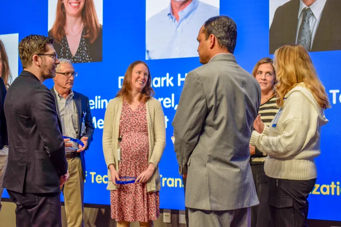 6 people stand mingling. Some hold the blue glass award they received. 