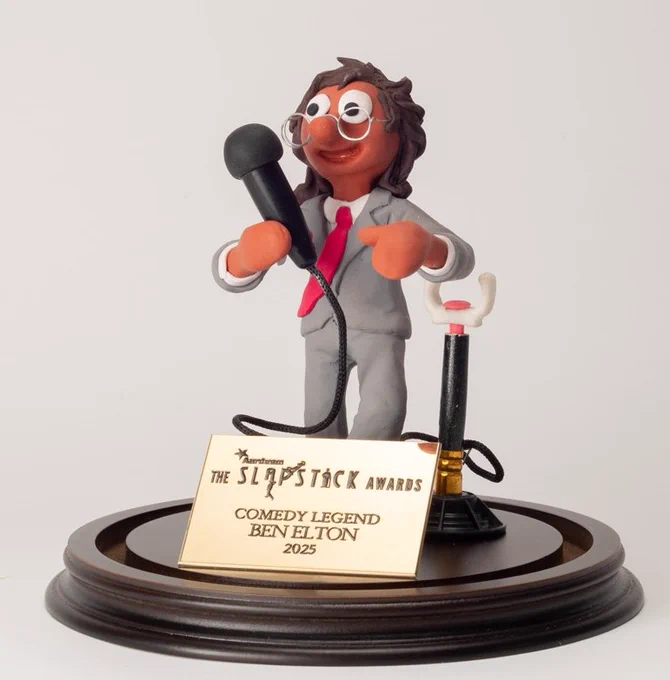 The Aardman Slapstick Comedy Legend award outside of the glass case, a morph dressed like Ben Elton doing stand-up (with microphone and microphone stand, wearing a grey suit and red tie) on a black plinth and plaque with "The Slapstick Awards - Comedy Legend Ben Elton 2025" engraved.