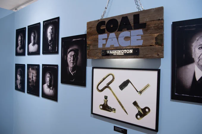 A selection of black and white tintype portraits on a pale blue painted wall. In the centre of the portraits is a painted wooden sign which reads 'Coal Face, Washington Co Durham'.