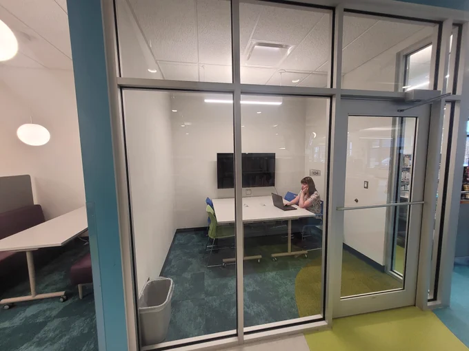 A white woman studies on her laptop in a closed, glass room. 
