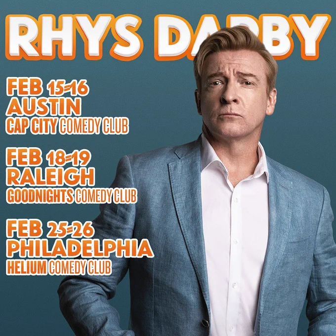 Rhys darby stands in front of a blueish green background in a blue suit with white undershirt looking serious and handsome, or seriously handsome, however you'd like to look at it. Text in Orange says: Rhys Darby Feb 15-16 Austin Cap City Comedy Club, Feb 18-19 Raleigh Goodnights Comedy Club - Feb 25-26 Philadelphia Helium Comedy Club