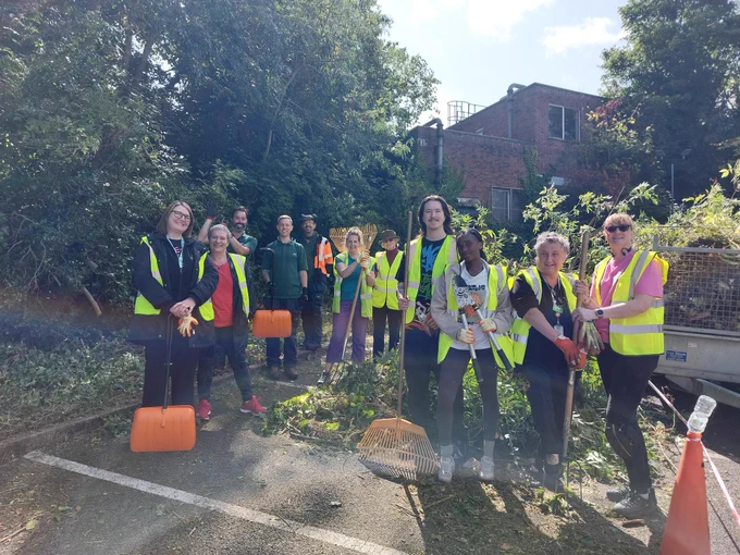 A group of 11 volunteers in hi-vis vests stand smiling and holding gardening equipment in a car park, next to a pile of leaves and tree cuttings