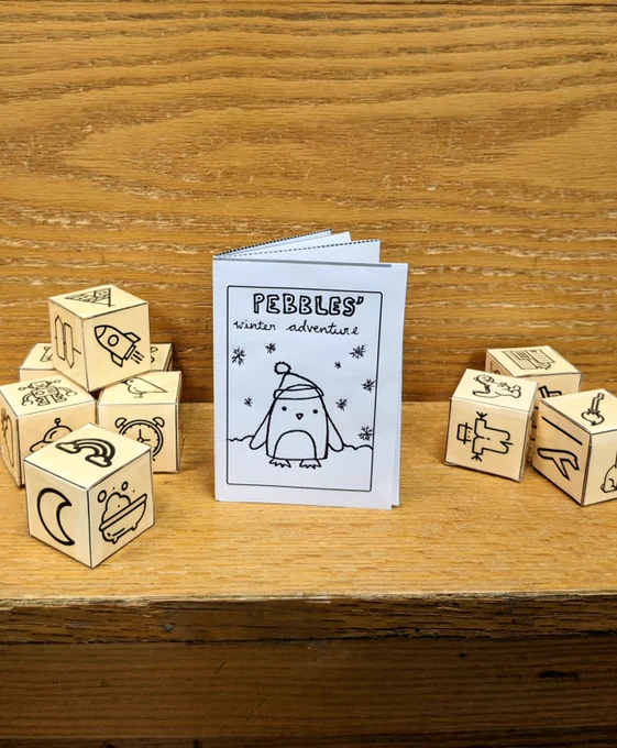 Photo of wooden shelf with a small story book template titled "Pebble's winter adventure", surrounded by a couple of piles of small cubes folded from paper with symbols on each side, including a crescent moon, a rocket, an alarm clock, a plane, a dinosaur and a tent.