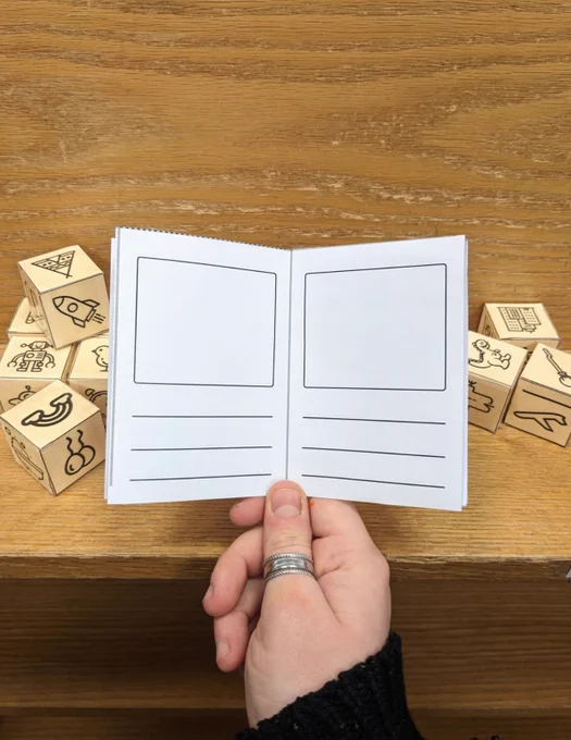 Photo of hand with a ring holding a storybook template open on a double page spread, in front of a pile of small cubes folded from paper with symbols on each side, including a rainbow,  balloons, a robot, a tent, a dinosaur and a plane.