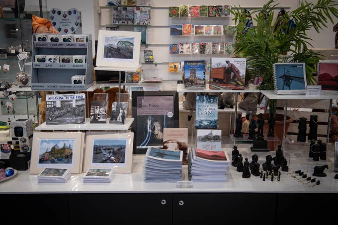 A display of products inside a brightly lit shop. On the shelves are a selection of books, colourful prints and different sized figures of coal miners.