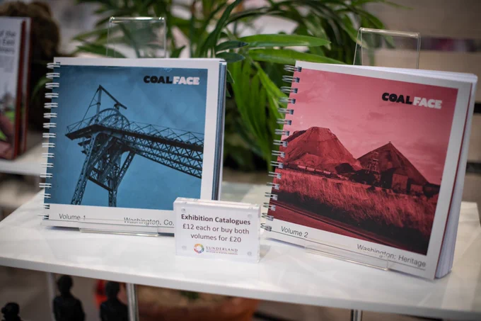 Two exhibition catalogues displayed on a white shelf. One had a blue front cover and the other is red. There is a sign which reads 'Exhibition Catalogues, £12 each or buy both for £20'.
