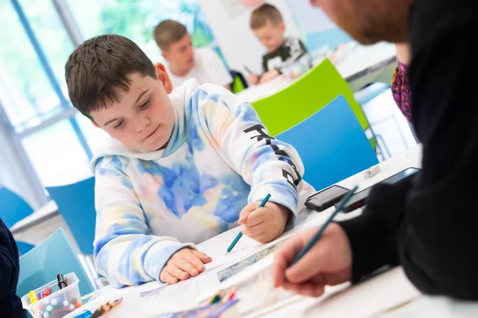 A child with short brown hair wearing a blue, pink and white tie due hoodie. They are sat at a table and are drawing on a piece of paper with a coloured pencil.