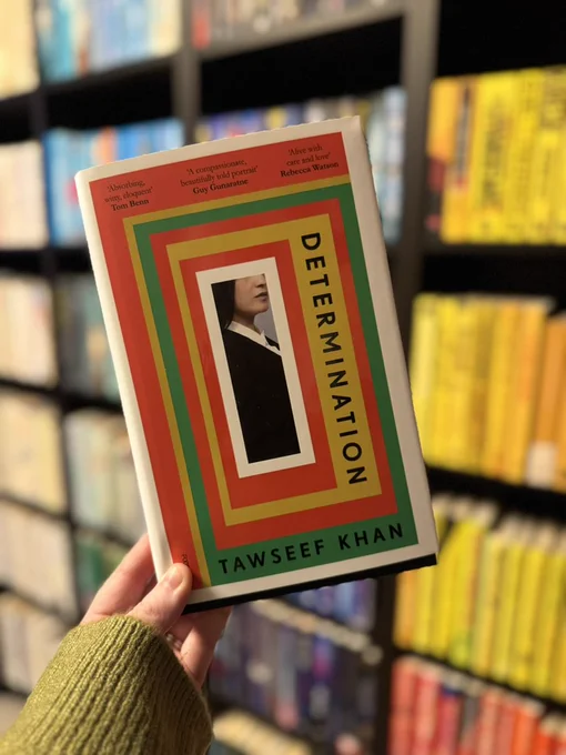 Determination (Hardback) by Tawseef Khan held up in front of a book case filled with books. 