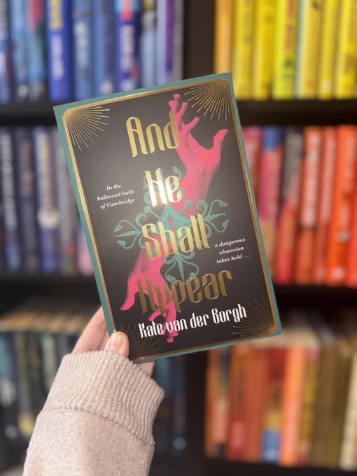 And He Shall Appear by Kate Van Der Borgh held up in front of a book case filled with books. 