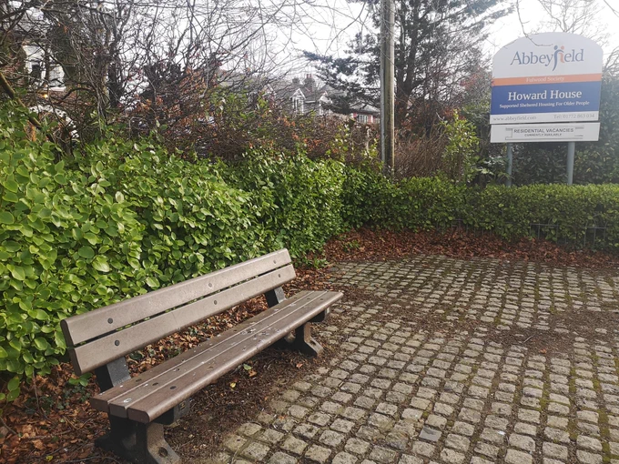 Bench with bird poo making it unusable. The bench is set on cobbles and is surrounded by green shrubbery. Next to Howard House residential home.