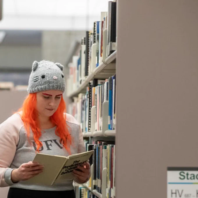 A woman stands near a library shelf and reads a book. She wears a took and UFV hoodie.