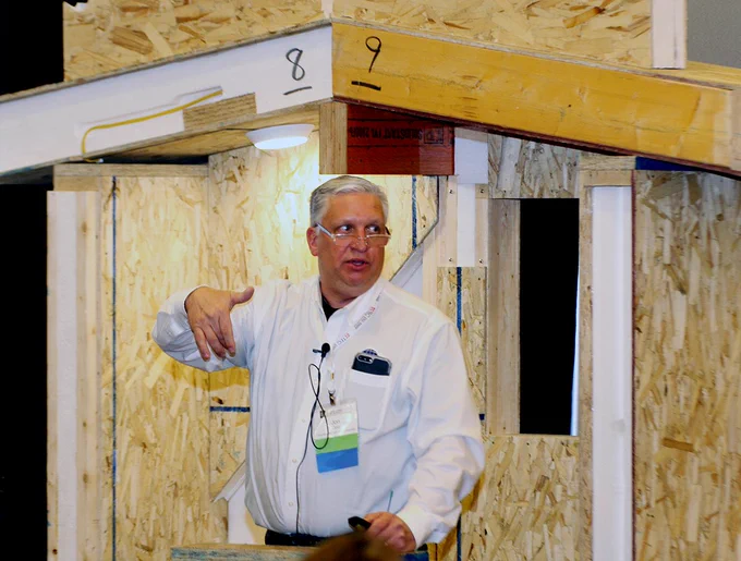 Person presenting at a construction trade show, standing in front of a wooden frame structure, wearing a badge and giving a thumbs-up.