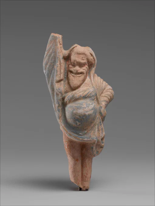 Photo of a small statue of a figure with a comedy face mask and a beard and a big belly. The figure has one arm raised and the other hand on their hip, and faded blue paint has chipped off to show reddish material underneath.
