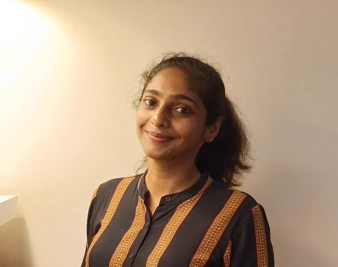The image shows Blessy smiling gently, with her dark hair tied back in a ponytail. She is wearing a dark-colored shirt with vertical orange and yellow stripes. The background is a plain, light-colored wall, softly illuminated by a light source in the top left corner, creating a warm and inviting atmosphere. In list,