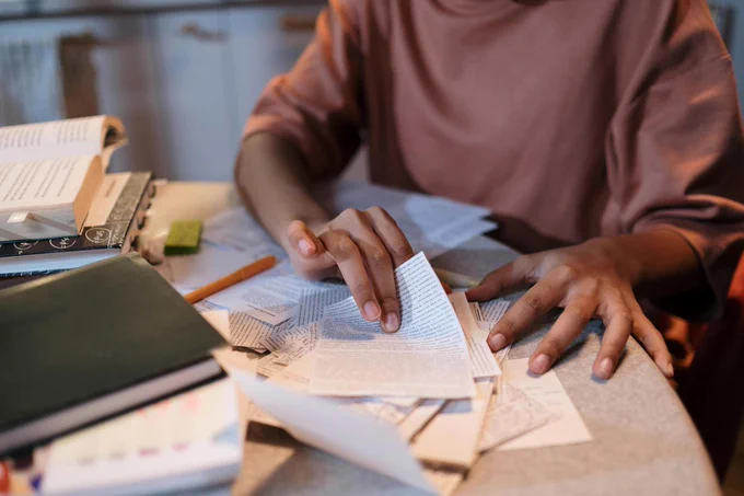 A person sitting at a desk with book and cut-outs of papers of text with various other papers.