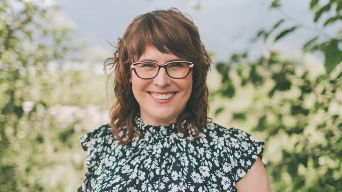 Smiling individual wearing glasses and a floral top, standing outdoors with green foliage in the background.