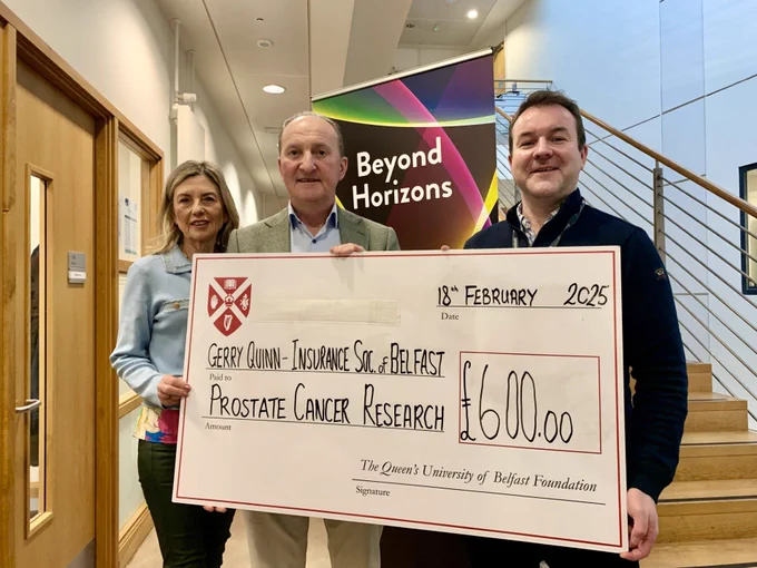 Photograph. Left to right: Geraldine Quinn, Gerry Quinn & Aidan Cole standing at the bottom of the PGJCCR stairs holding a large cheque that reads: Gerry Quinn - Insurance Society of Belfast, made out to Prostate Cancer Research via Queen's University of Belfast Foundation for £600.
