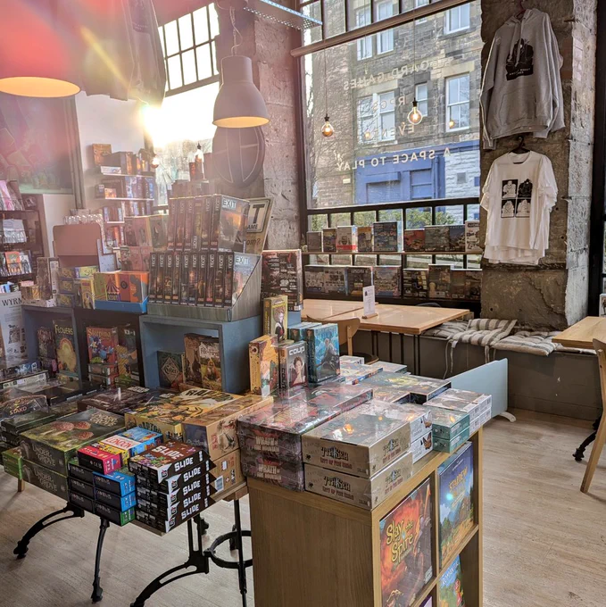 Interior of the board game shop in Edinburgh Ancient Robot Games. There is a merch table full of board games at the forefront. Light is coming in from the large windows behind the table.