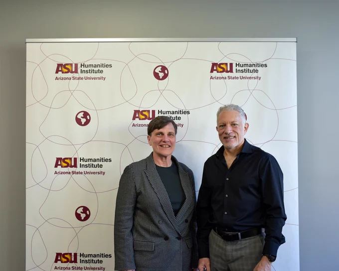 Paula Krebs and Ron Broglio smile together in front of a step and repeat (banner), which has the ASU Humanities Institute logo patterned throughout it.