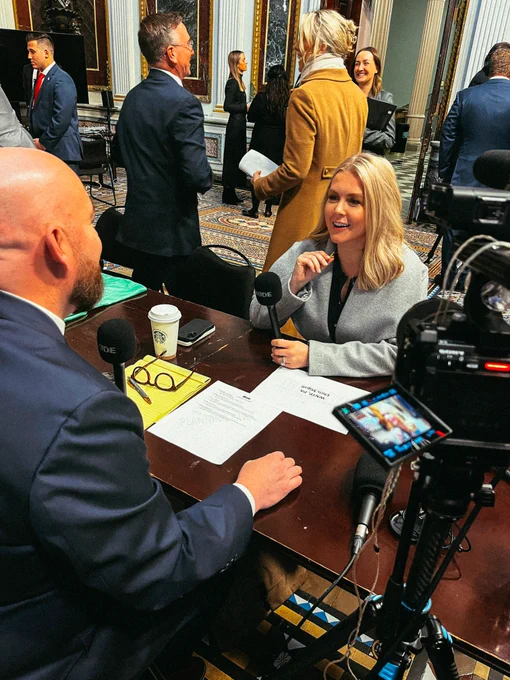 A vibrant shot of Karoline Leavitt speaking to Chris Stigall at the White House during a media row event. Karoline is confidently addressing a group of journalists, with the White House visible in the background. The atmosphere is energetic, with various media outlets gathered around, capturing the moment. The image conveys a sense of leadership and progress, highlighting Karoline’s role in connecting the White House to the nation.
