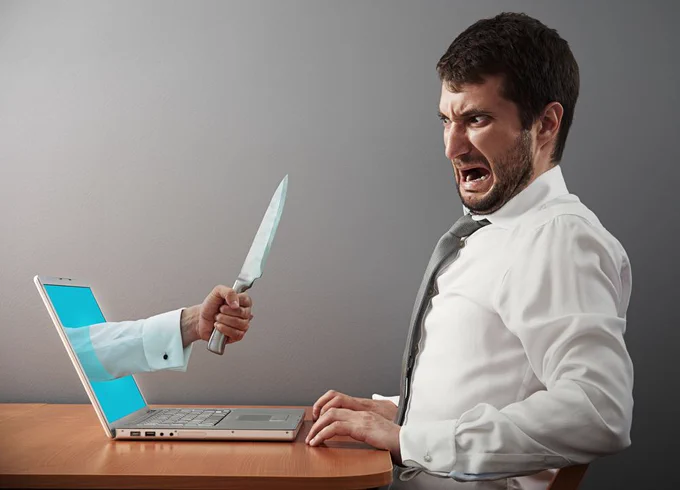 A man in a shirt and tie sits at a laptop looking horrified. From the screen, an arm appears holding a knife aggressively.