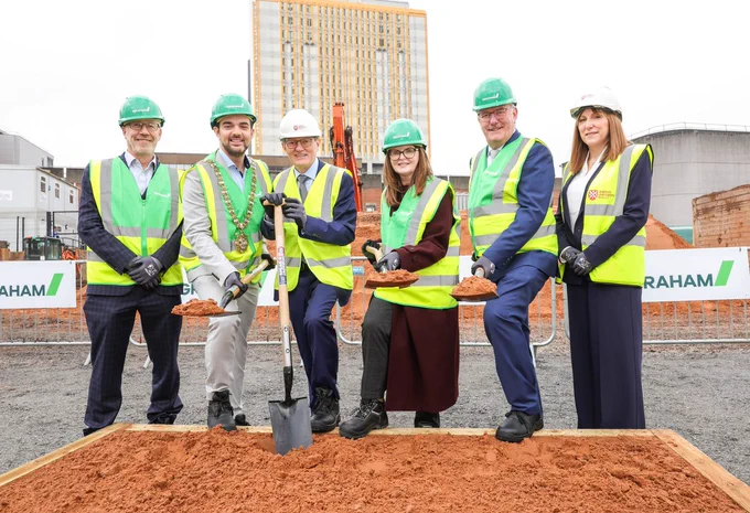 Image shows Professor Sir Ian Greer, President and Vice-Chancellor of Queen's University; Professor Judy Bradley, Director of iREACH Health; Economy Minister Dr Caoimhe Archibald; Health Minister Mike Nesbitt; Professor Ian Young, Chief Scientific Advisor to the Northern Ireland Department of Health; Councillor Micky Murray, Lord Mayor of Belfast, pictured at the sod-cut. Professor Sir Ian Greer is holding a shovel and the group are all wearing hi-vis vests and hard hats.