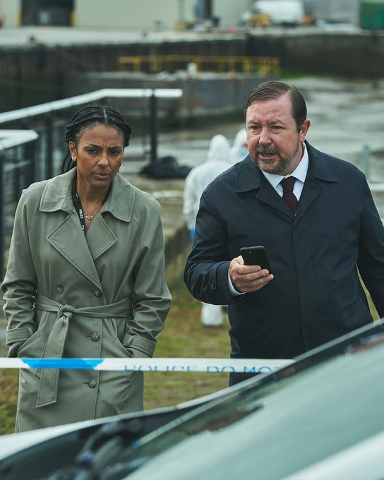 Two detectives, one in a green trench coat and the other in a navy overcoat, walk past police tape. Behind them, forensic investigators in white suits examine the area near a dock.
