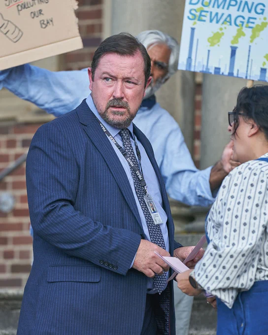 A detective in a pinstripe suit stands amid a tense protest, holding papers while looking concerned. Behind him, demonstrators hold signs about pollution and sewage. 