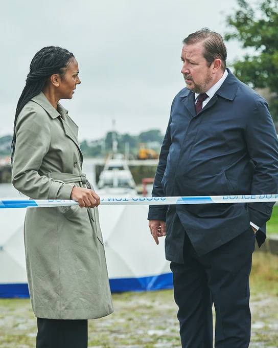 Two detectives stand by some police tape, engaged in a serious conversation at a crime scene near the water. A forensic tent is visible in the background. 