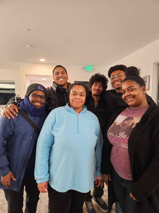 A group of six people standing together indoors, dressed in winter clothing.